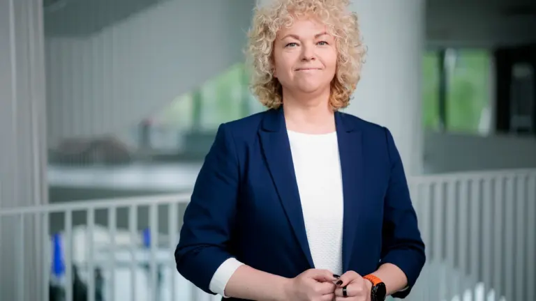 Šviesiaplaukė garbanota moteris vilkinti tamsiai mėlyną švarką stovi modernioje šviesioje biuro aplinkoje.