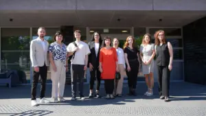 Devyni įvairaus amžiaus žmonės stovi kartu po stogeliu prie šiuolaikinio pastato įėjimo.