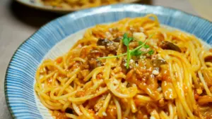 Spageti makaronai su raudonu padažu, grybais ir šviežiais žalumynais dryžuotame mėlynos spalvos dubenyje.
