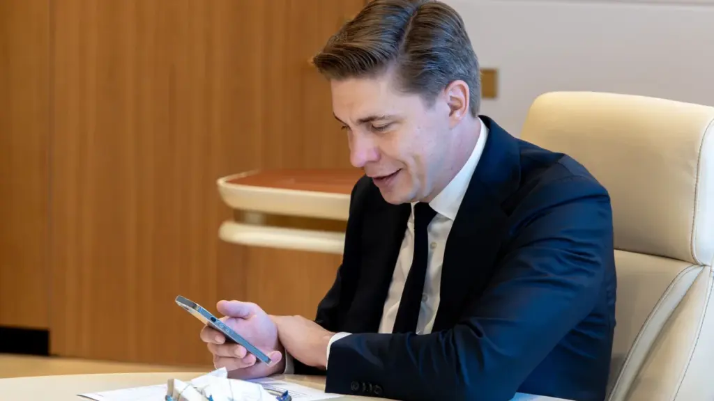 Šviesiaplaukis vyras tamsiu kostiumu sėdi prie stalo ir žiūri į išmanųjį telefoną.