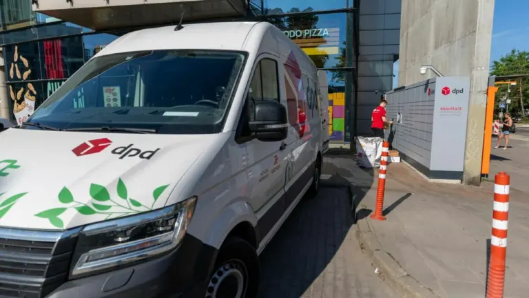 Baltas DPD kurjerių furgonas stovi šalia DPD siuntų terminalo su dirbančiu darbuotoju.