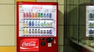 Ryškiai raudonas „Coca-Cola“ gėrimų automatas šviečia šiuolaikinėje miesto aplinkoje.