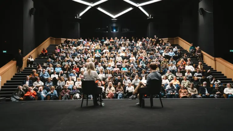 Du žmonės sėdi scenoje ir kalbasi pilnoje modernioje teatro salėje prieš žiūrovų auditoriją.