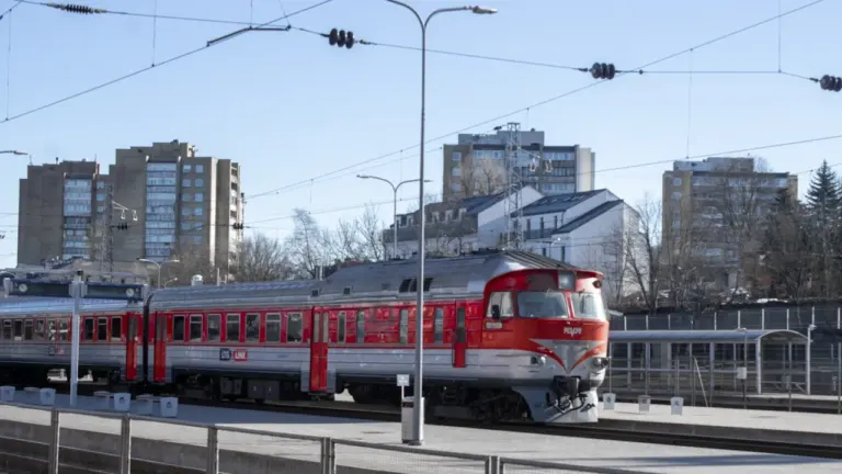 Raudonas traukinys rieda pro Vilniaus stotį saulėtą dieną, fone matomi daugiabučiai.