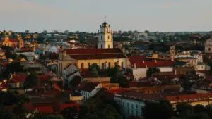 Panoraminis Vilniaus senamiesčio vaizdas su raudonais stogais ir bažnyčios bokštu saulėlydžio metu.