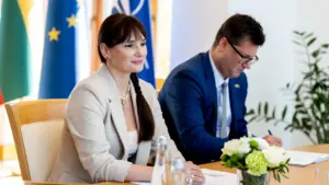 Moteris ir vyras sėdi prie stalo priešais vėliavas oficialiame politiniame susitikime.
