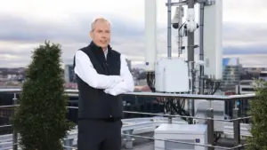 Vidutinio amžiaus vyras stovi ant pastato stogo šalia ryšio antenų bokšto miesto fone.