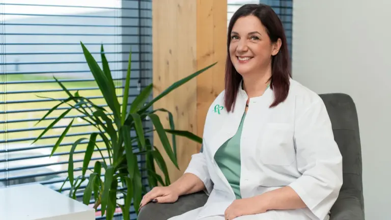 Besišypsanti tamsiaplaukė gydytoja balta medicinine apranga sėdi fotelyje šalia žaliuojančio kambarinio augalo.