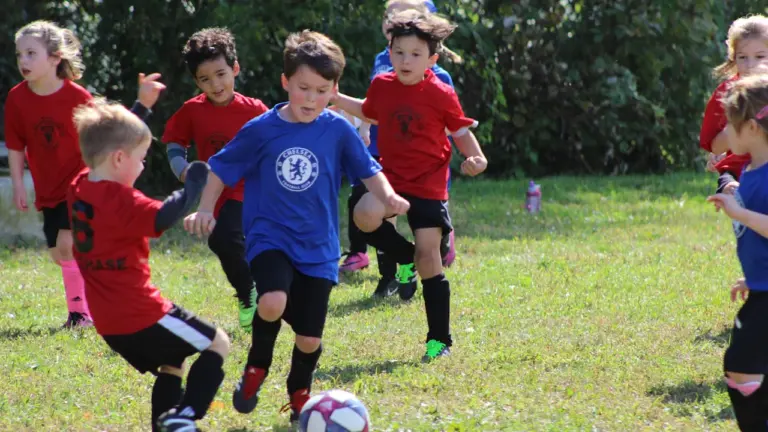 Vaikai su sportine apranga žaidžia futbolą lauko aikštelėje vasaros dieną