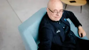 Plikai skustas vyras su akiniais sėdi melsvame fotelyje ir žiūri tiesiai į fotoobjektyvą.