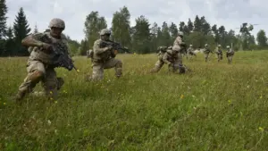 Kariai kamufliažinėmis uniformomis su ginklais lauke, fone matyti skrendantis karinis sraigtasparnis ir miškas.