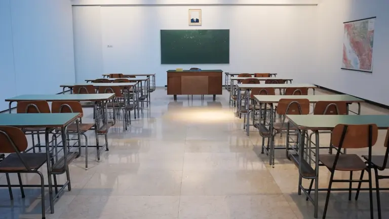 Tuščia klasė su mediniais stalais ir lenta, simbolizuojanti švietimo sistemos pokyčius.