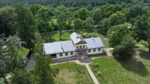 Vaizdas iš viršaus į restauruotą tradicinį Lietuvos dvarą, apsuptą žaliuojančio miško ir parko erdvės.