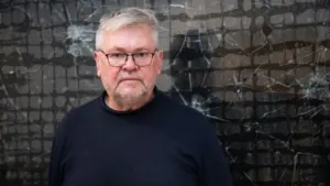 Vyresnio amžiaus vyras su akiniais stovi prieš tamsų, dūžusį lango stiklą su tinkleliu.