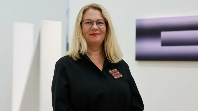 Šviesiaplaukė moteris su akiniais ir juodais marškiniais stovi šviesiame moderniame fone su meno kūriniais.