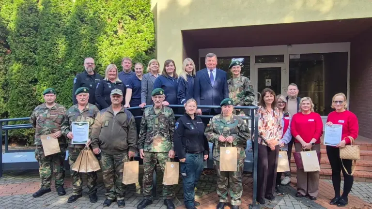 Didelė žmonių grupė su uniformomis ir civiliais drabužiais stovi prie pastato lauke laikančių padėkos raštus.