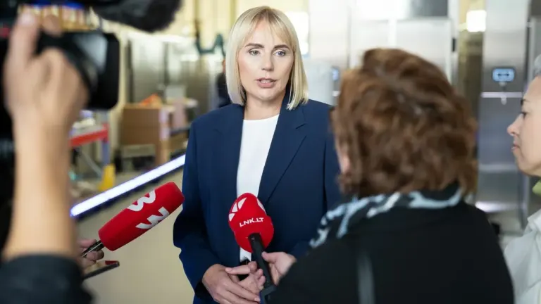 Šviesiaplaukė moteris tamsiai mėlynu švarku kalba į žurnalistų laikomus mikrofonus gamybinėse patalpose.