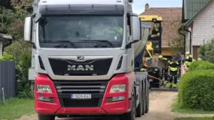 Pilkas sunkvežimis „MAN“ su šviesų signalu stovi gatvėje šalia kelio tiesimo technikos ir darbininkų.