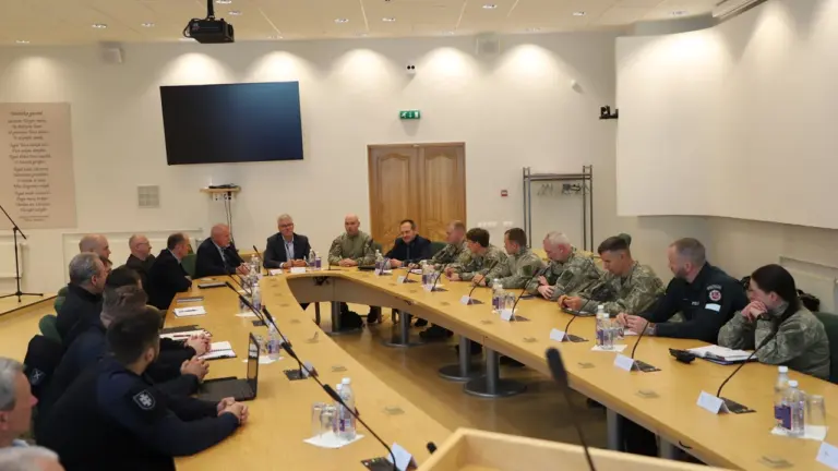 Karininkai ir pareigūnai sėdi prie didelio ovalaus stalo per oficialų posėdį šviesioje salėje.