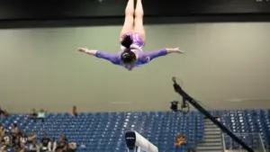 Sportininkė atlieka profesionalų aerobinės gimnastikos šuolį didelėje sporto arenoje prieš žiūrovų tribūnas.
