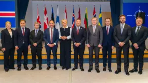 Grupė oficialiai apsirengusių politikų stovi rikiuotėje priešais įvairių valstybių vėliavas uždaroje patalpoje.