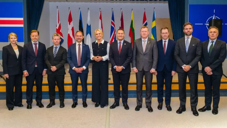 Grupė oficialiai apsirengusių politikų stovi rikiuotėje priešais įvairių valstybių vėliavas uždaroje patalpoje.
