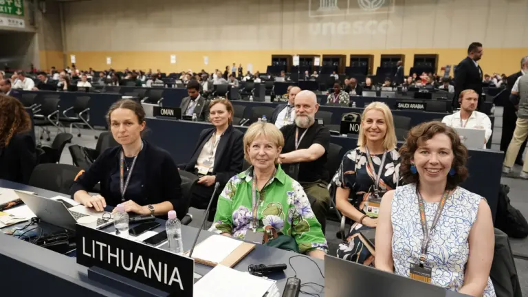 Lietuvos delegacija sėdi prie stalo tarptautinėje UNESCO konferencijoje su kompiuteriais ir užrašais.