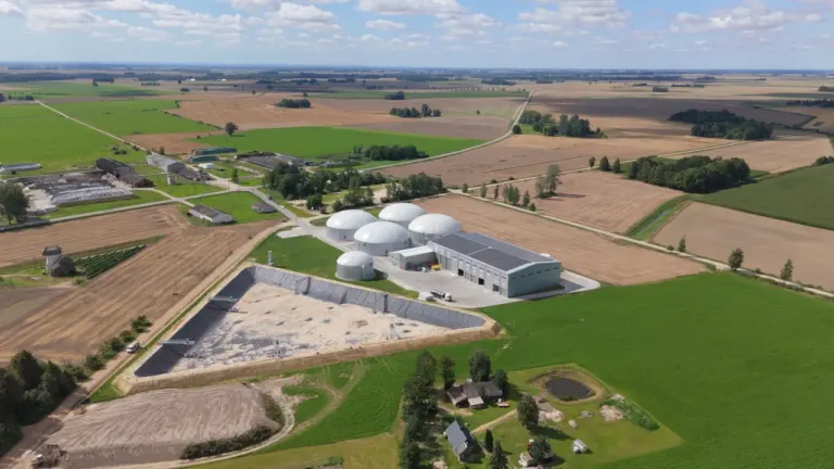 Iš oro matoma biodujų jėgainė su apvaliais kupolais apsupta plačių žemės ūkio laukų.