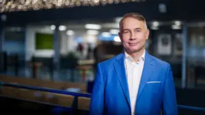 Vidutinio amžiaus vyras su mėlynu švarku ir baltais marškiniais stovi šiuolaikiškoje biuro patalpoje.