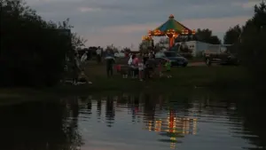 Spalvinga apšviesta karuselė stovi šalia ežero, kur žmonės leidžia laisvalaikį vakaro metu.